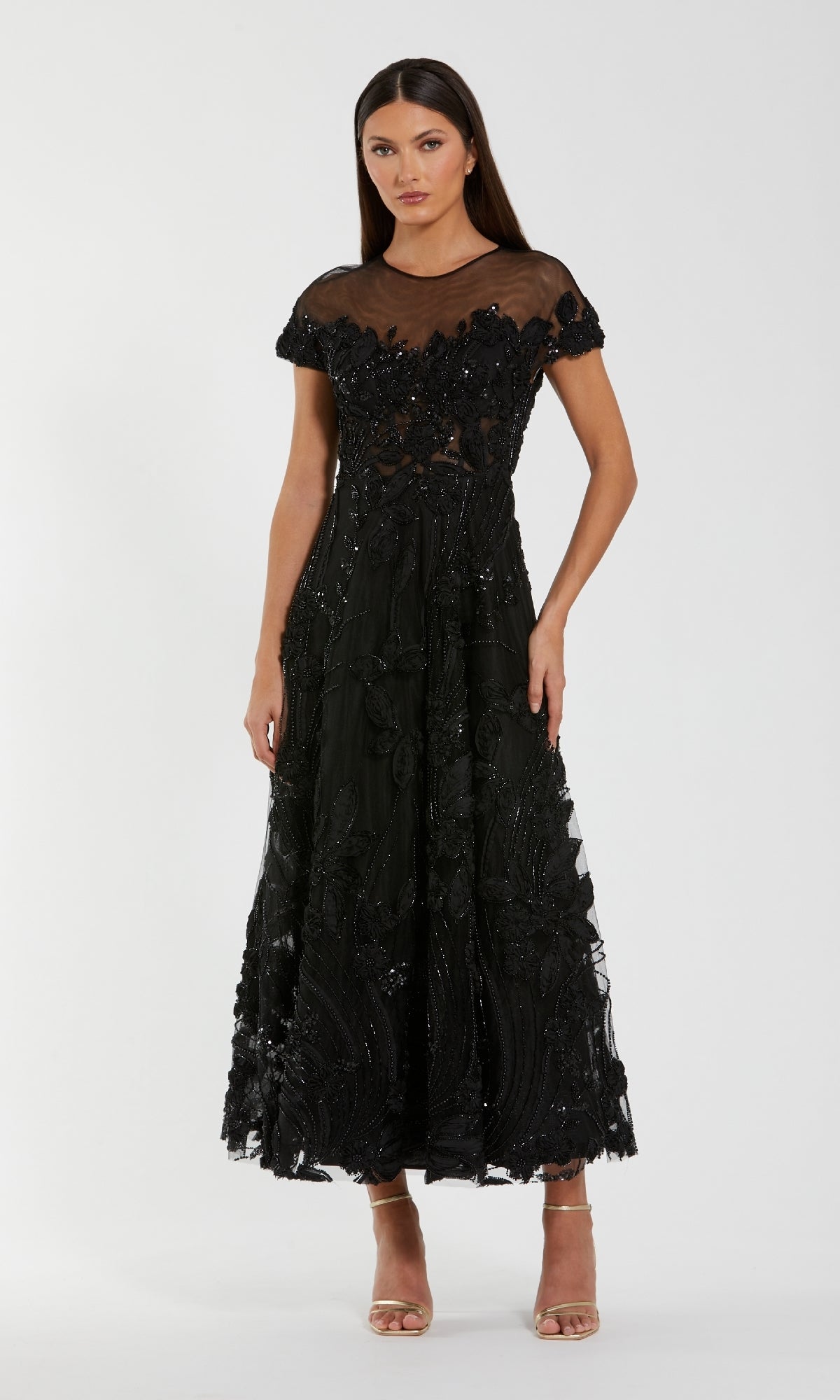 A woman wears the Mac Duggal 69003 Cap-Sleeve Black Formal Dress, a mid-length formal gown with sheer details and floral embellishments, styled with nude open-toe heels.